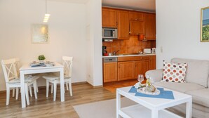 Une cuisine moderne avec des meubles en bois, un four à micro-ondes et une machine à café. On y trouve un coin repas avec une table et des chaises blanches, ainsi qu’un salon avec un canapé et une table basse.