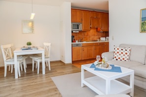 A modern kitchen with wooden cabinets, a microwave, and a coffee maker. There is a dining area with a white table and chairs, and a living room with a sofa and a coffee table.