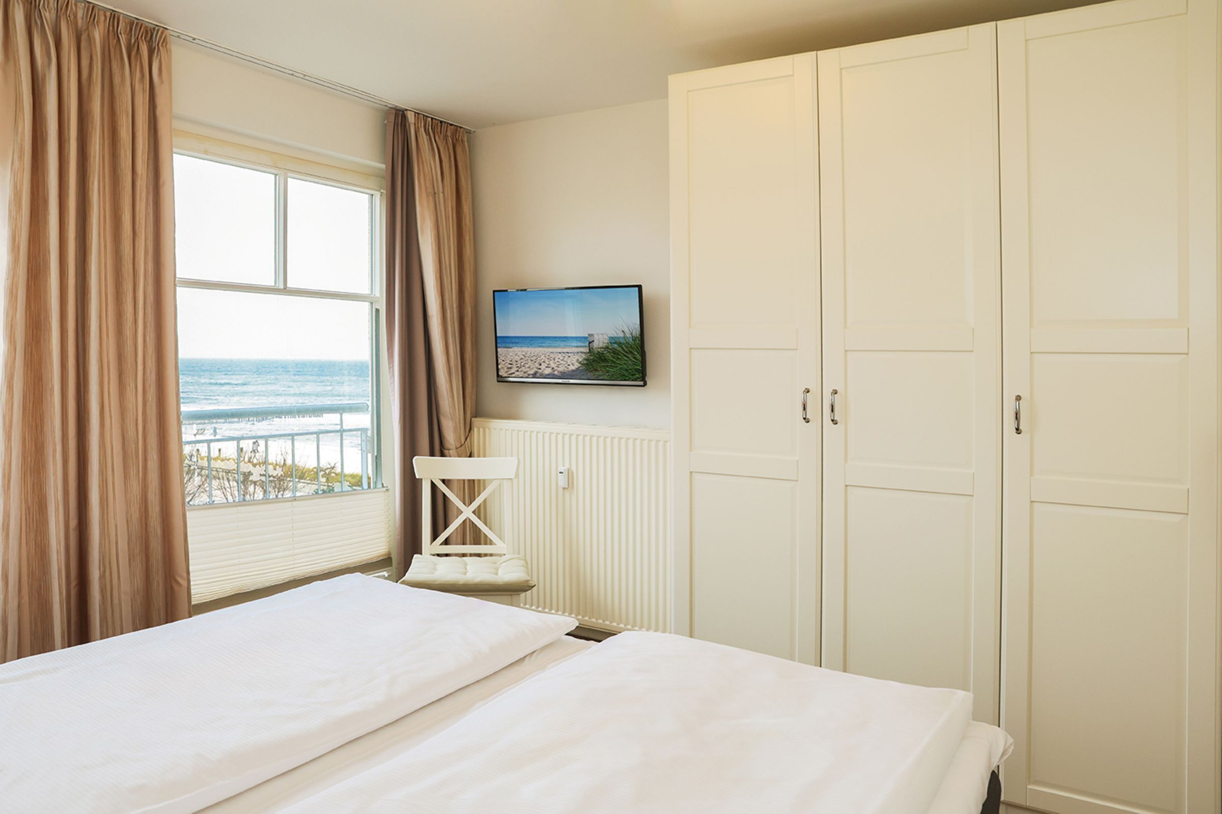 A bedroom with a bed, a window with curtains, a radiator, a television on the wall, and a wardrobe.