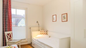 Une chambre avec un lit simple, une chaise, une table avec un jeu de société, une fenêtre avec des rideaux et deux tableaux encadrés accrochés au mur.