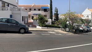 Una calle con coches aparcados, un edificio blanco con toldo azul y un árbol alto.