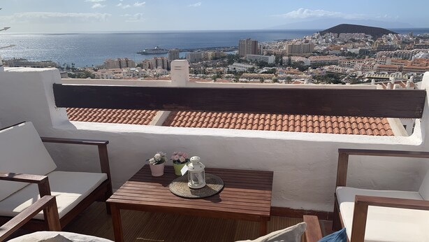 Una terraza con vistas al mar y a la ciudad, que incluye una mesa de madera, sillas y un área de descanso acolchada.