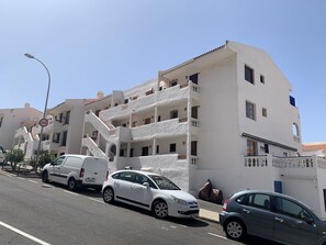 A multi-story building with balconies, a street with parked cars, and a speed limit sign.