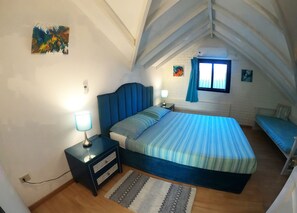 A bedroom with a large bed, a nightstand, a lamp, a power outlet, a window with curtains, and a small wall art piece.