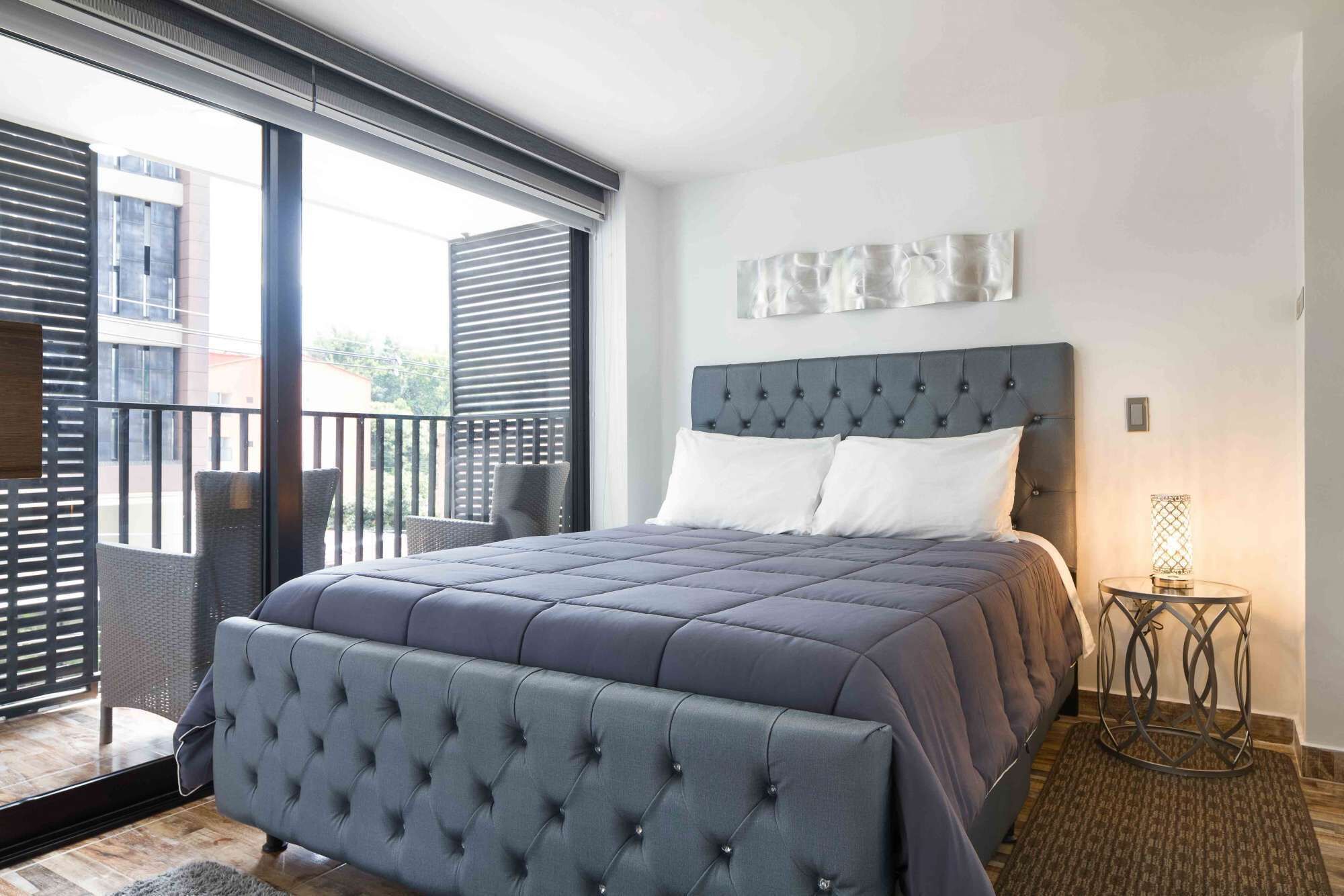 A bedroom with a large bed, a headboard, a nightstand, and a view of the outside.