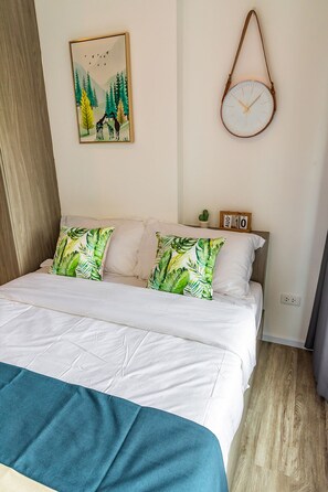 A neatly made bed with white bedding and two green leaf-patterned pillows, a wall-mounted clock, and a framed nature painting.