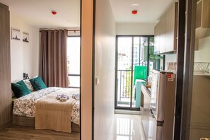 A bedroom with a bed, a window with curtains, a balcony, and a kitchen area with a refrigerator.