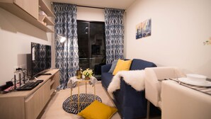 Een moderne woonkamer met een blauwe bank, een witte fauteuil, een klein rond tafeltje met een lamp, en een televisie op een houten dressoir.