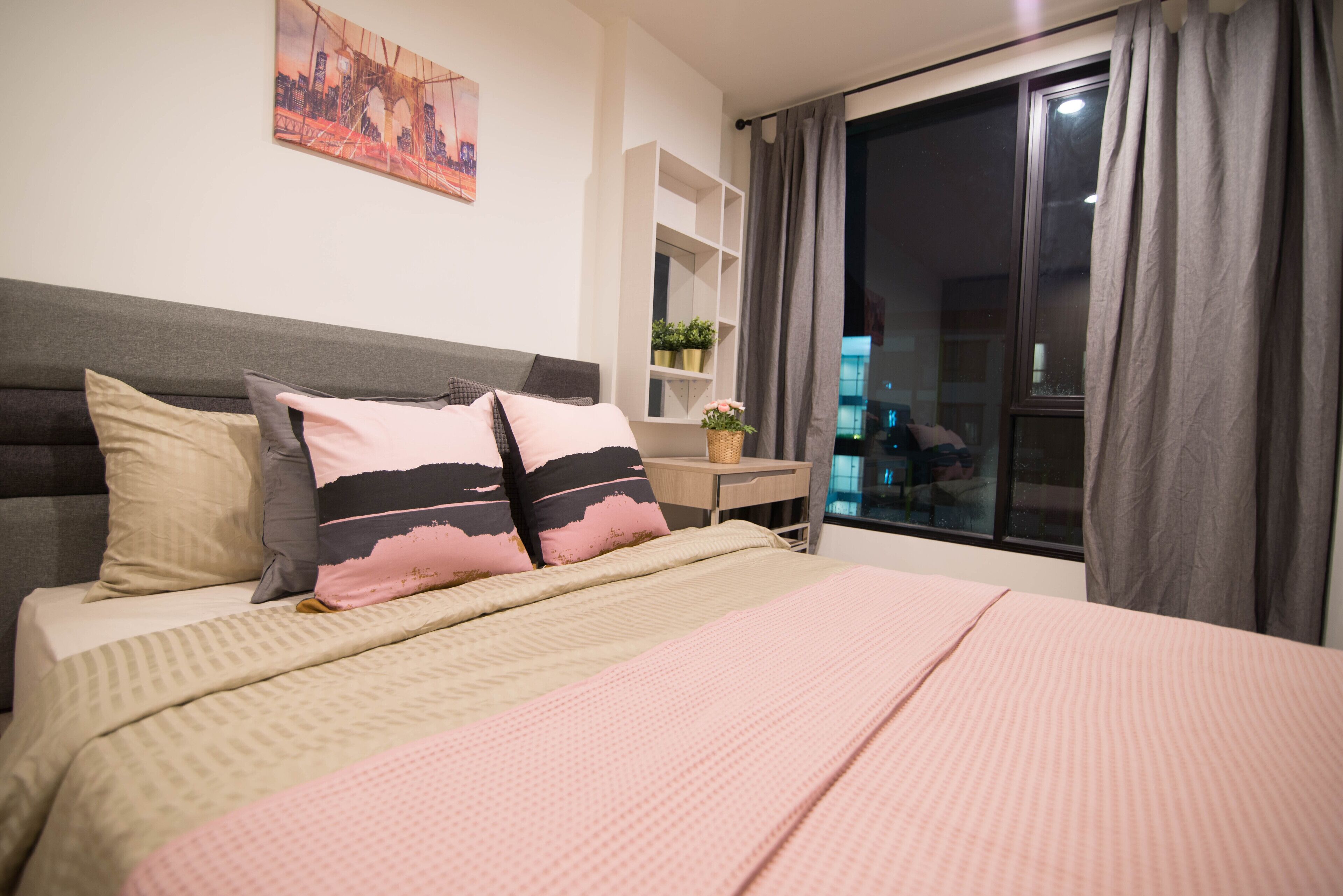 Cama bem arrumada com colcha rosa e bege, cabeceira com painel cinza e uma pintura de paisagem urbana na parede.