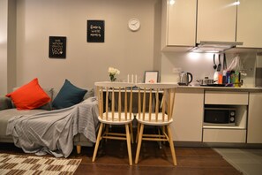 A compact living space with a sofa, two wooden chairs, a small table, a kitchen area with a microwave, and a clock on the wall.