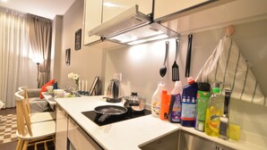 Eine moderne Küche mit weißer Arbeitsplatte, Spüle aus Edelstahl und diversen Küchengeräten. Auf der Arbeitsplatte stehen Reinigungsmittel, und im Hintergrund ist ein Stuhl zu sehen.