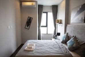 A neatly made bed with a white comforter and pillows, a bedside table with a lamp, and a window with curtains.