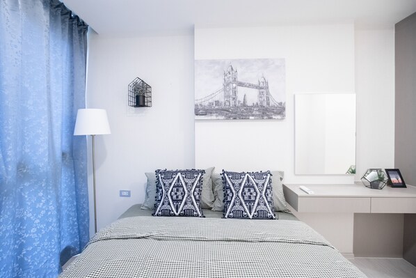 A bedroom with a bed, a desk, a lamp, a picture of the Tower Bridge, and a window with curtains.