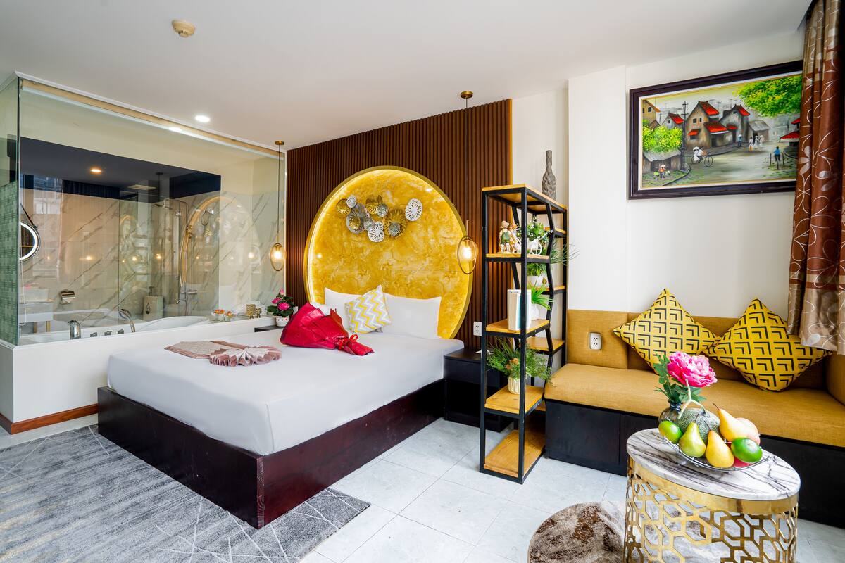A modern bedroom with a large bed, a glass-enclosed shower, a wooden headboard, a sofa with patterned cushions, and a small table with fruit.