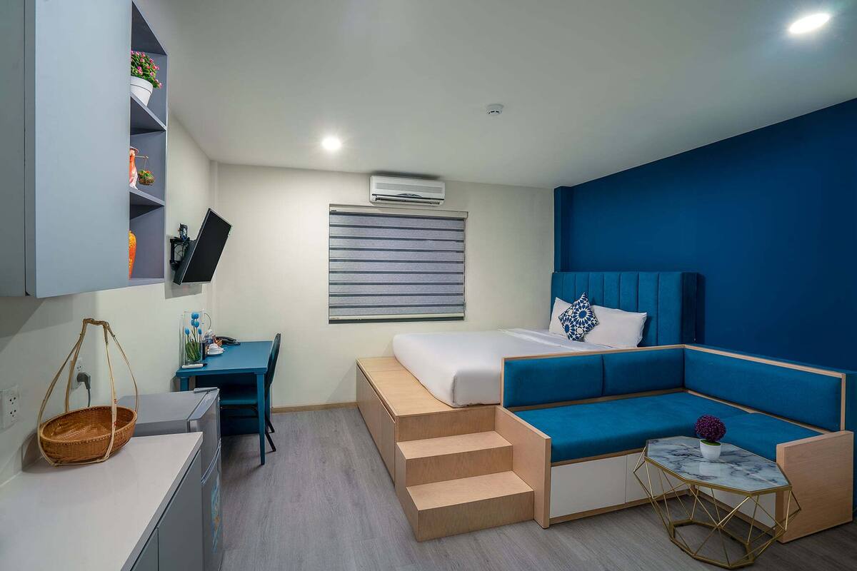 A compact hotel room with a bed, a desk, a chair, a small table, and a built-in shelf with decorative items.