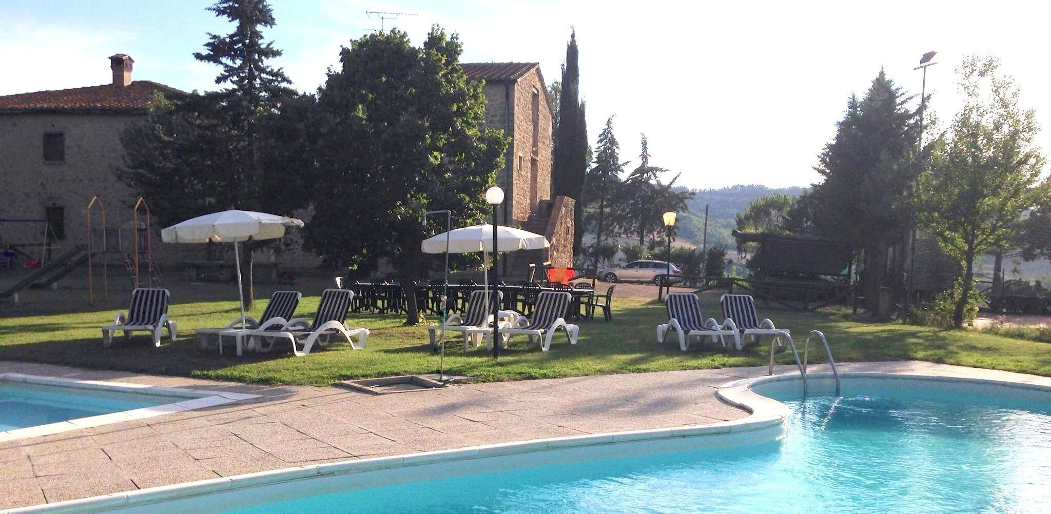 A pool area with lounge chairs and umbrellas, surrounded by trees and a building with a stone facade.
