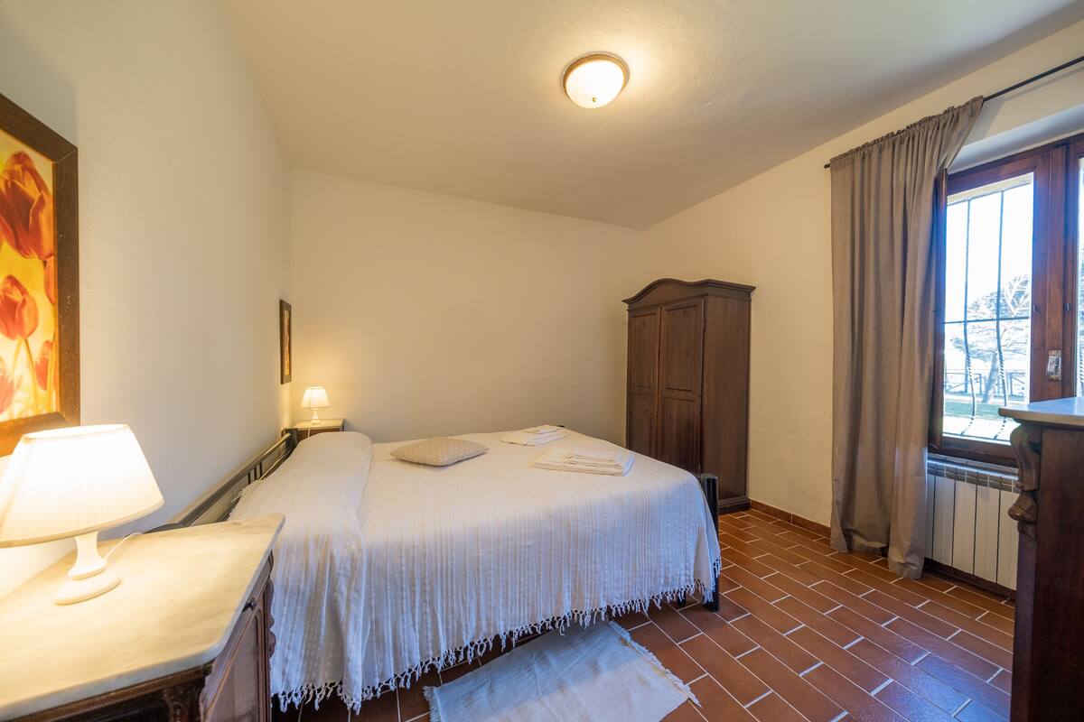 Une chambre à coucher comprenant un lit, une table de chevet, une lampe, une fenêtre avec des rideaux et un radiateur.