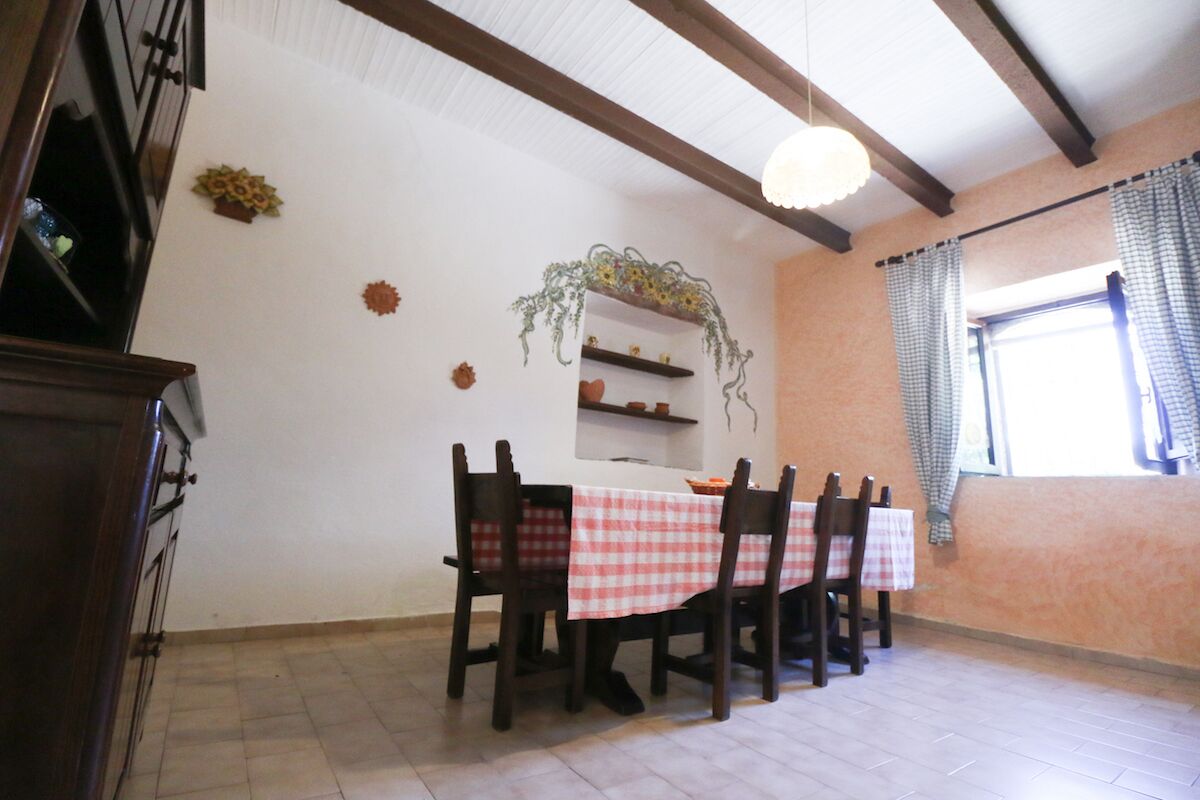 Un coin repas avec une table dressée pour quatre personnes, des chaises en bois, un mur blanc avec une étagère et une fenêtre avec des rideaux.