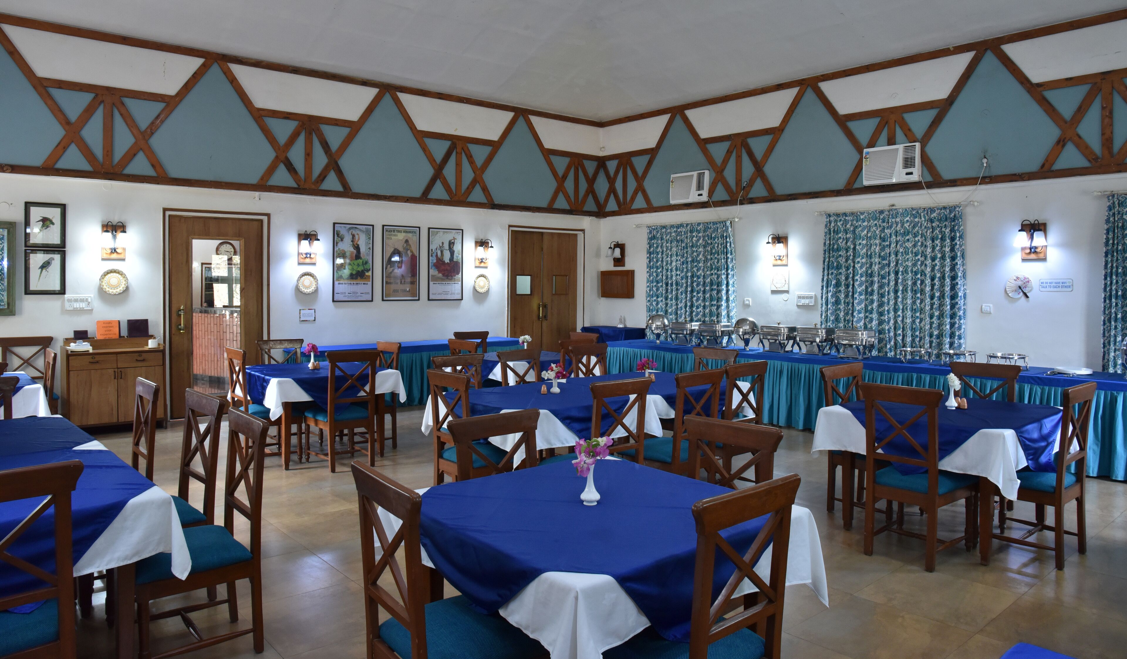 Un comedor con varias mesas cubiertas con manteles azules, sillas de madera y unidades de aire acondicionado fijadas a la pared.