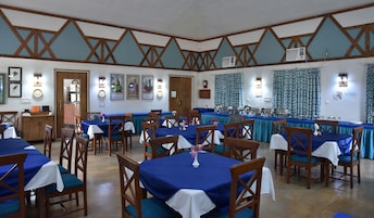 Un comedor con varias mesas cubiertas con manteles azules, sillas de madera y unidades de aire acondicionado fijadas a la pared.