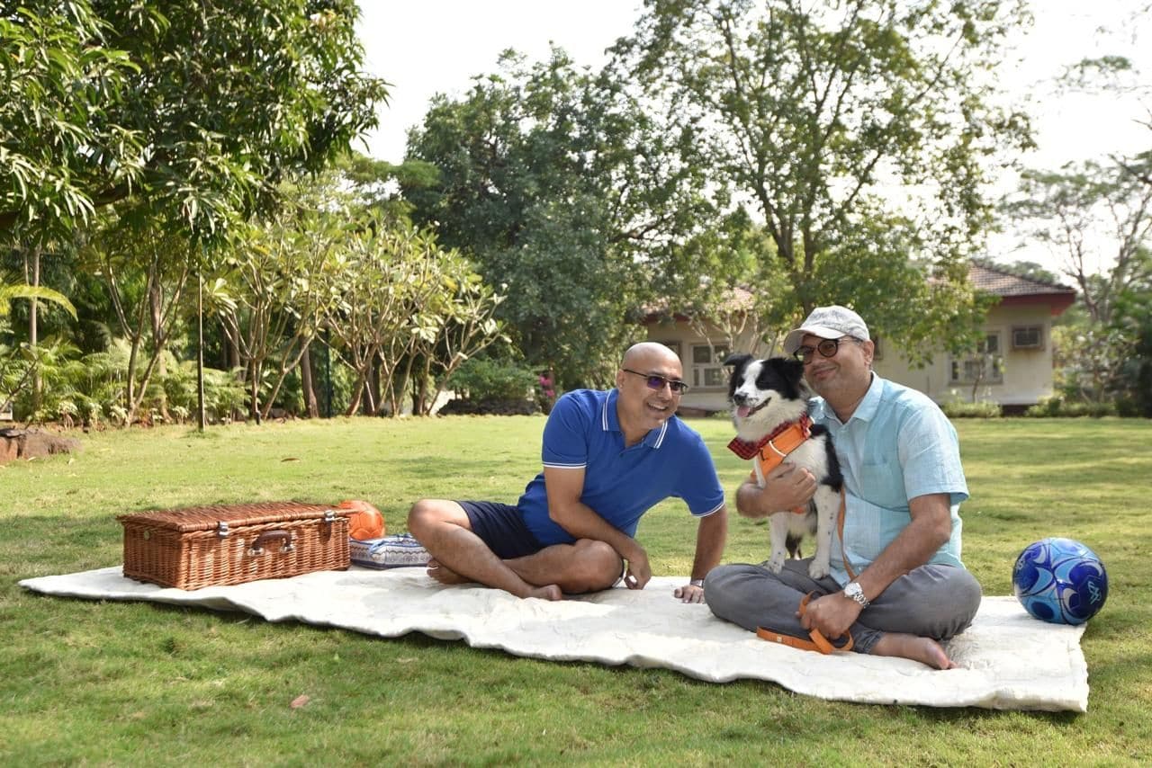 Deux hommes sont assis sur une couverture à l’extérieur, accompagnés d’un chien, d’un panier et d’un ballon de football.