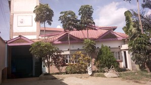 A two-story building with a red roof, surrounded by trees and a dirt driveway. - Palin Hotel (Mwingi)