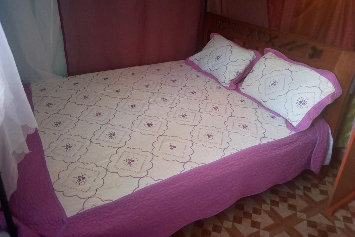 A single bed with a patterned bedspread and two pillows, a wooden headboard, and a checkered floor.