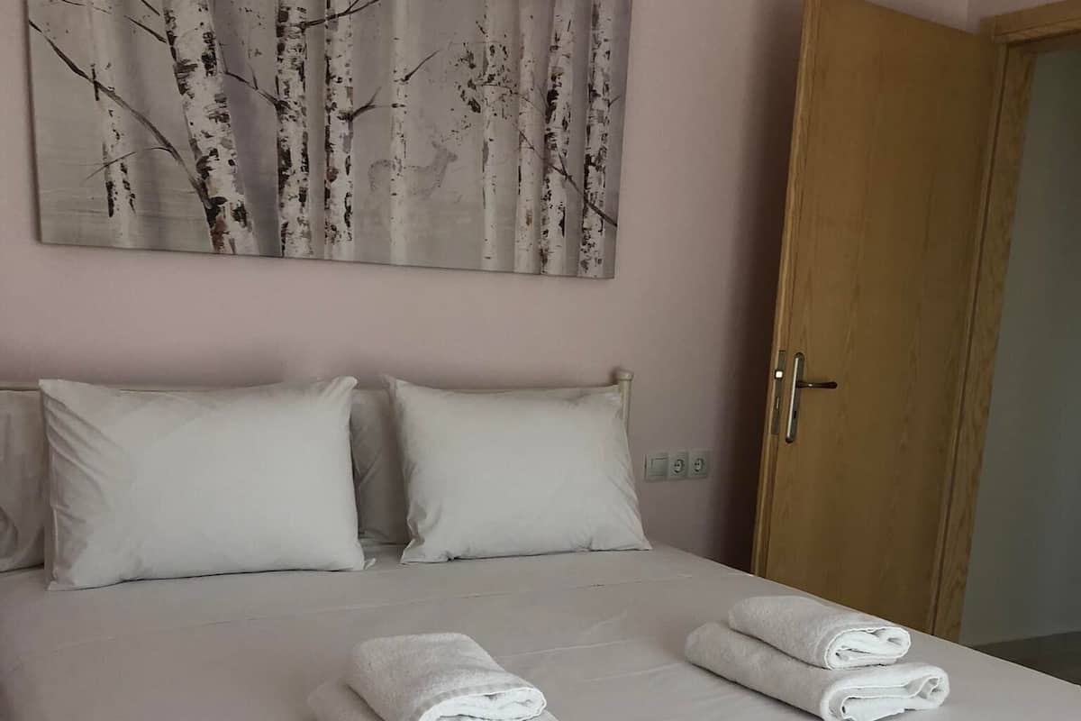 Cama bem arrumada com lençóis brancos e duas toalhas enroladas, uma porta de madeira e um quadro com paisagem de bétulas na parede.