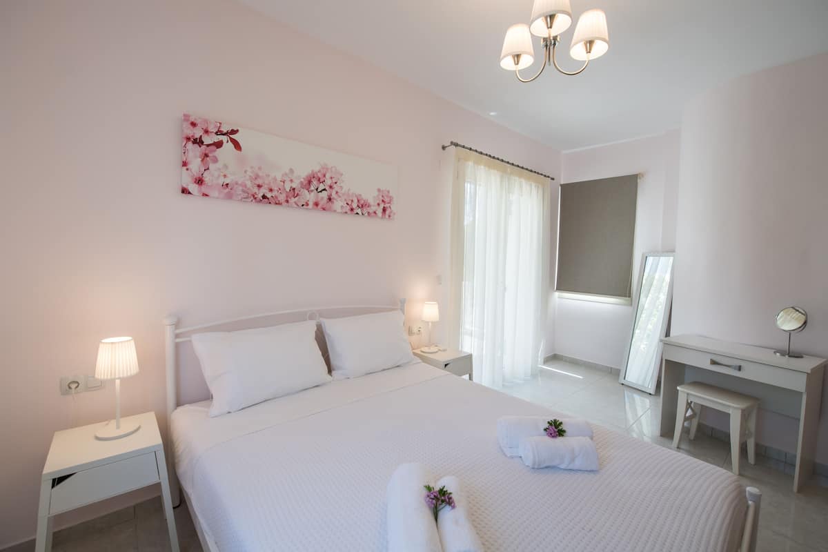 Quarto de hotel com cama, mesas de cabeceira, escrivaninha, cadeira, espelho e janela com cortinas.