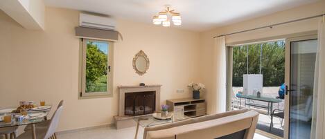 A modern living room with a glass coffee table, beige sofa, and a fireplace. There is a dining area with chairs and a table set for breakfast.