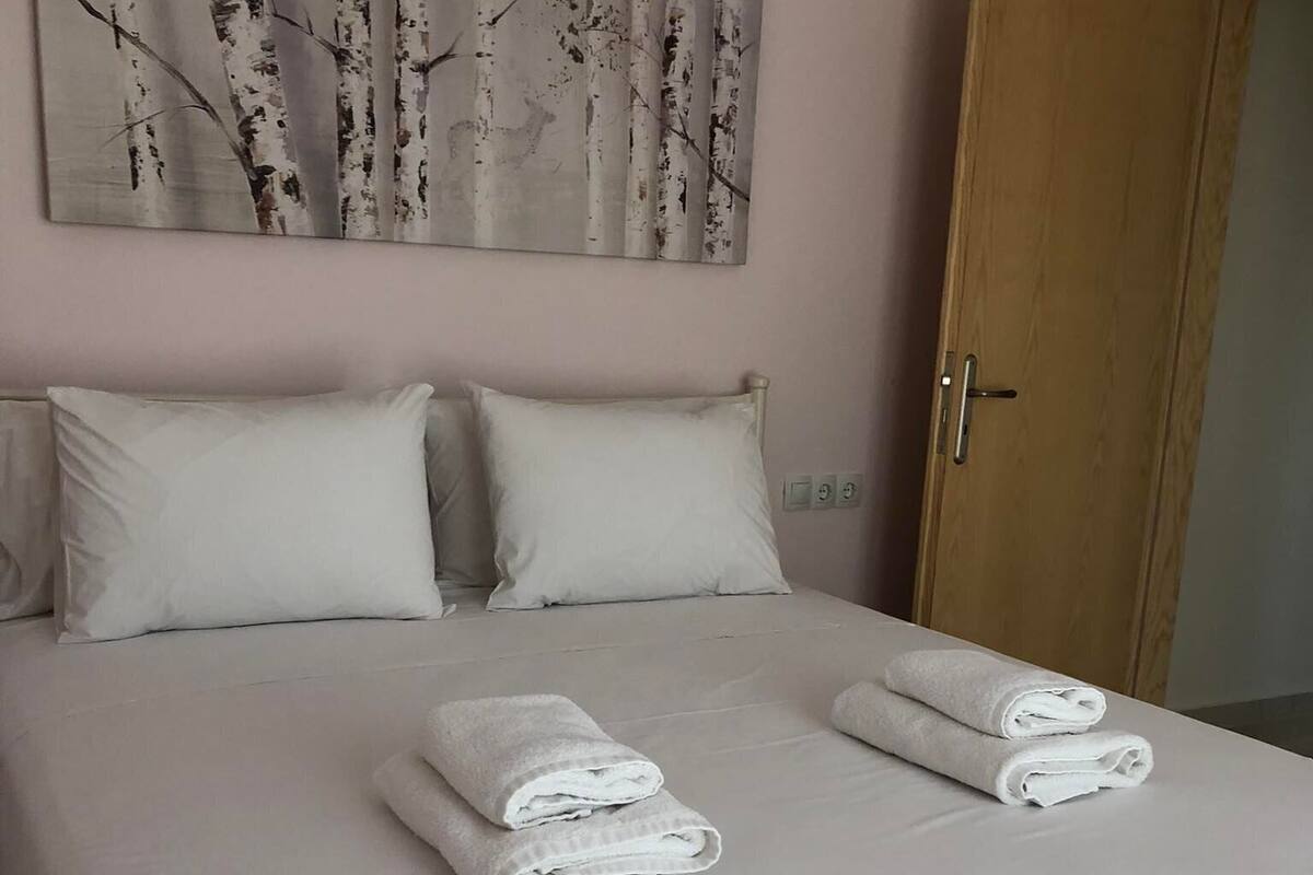 Uma cama bem arrumada com lençóis e toalhas brancas, uma porta de madeira e um quadro com árvores de bétula na parede.