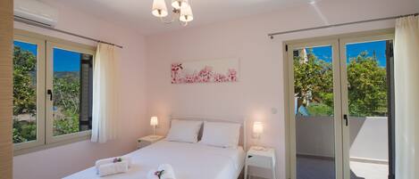 Une chambre à coucher comprenant un lit, deux tables de chevet, un lustre, une fenêtre avec des rideaux et une porte-fenêtre coulissante donnant sur un balcon.