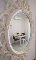 A room with a white ornate mirror reflecting a dining area with a table, chairs, and an air conditioning unit on the wall.