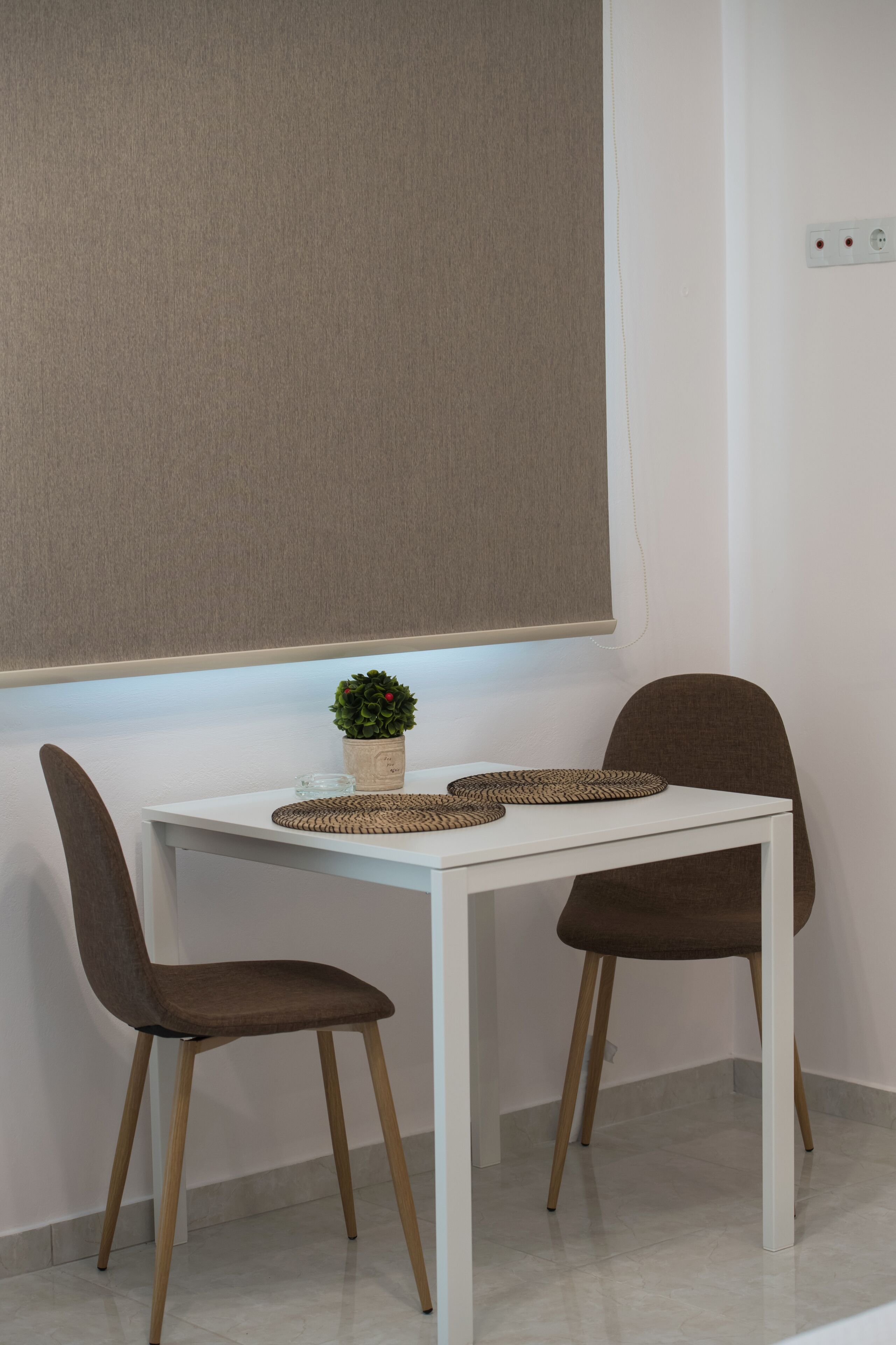 A white table with two brown chairs, a small potted plant, and a woven placemat.
