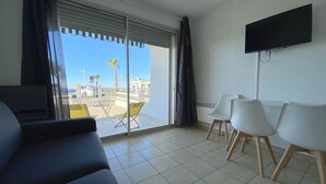 A room with a sofa, two chairs, a TV, and a large sliding glass door leading to a balcony with a view of palm trees and buildings. - Sol-y-Days le Goéland, Seafront apartment with parking (Le Grau-du-Roi)