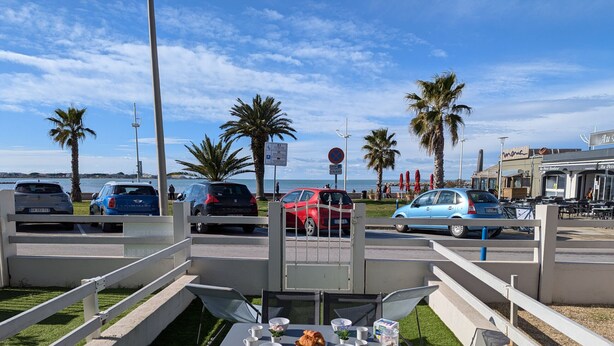 An outdoor seating area with tables and chairs, a view of the sea, and parked cars. - Sol-y-Days le Goéland, Seafront apartment with parking (Le Grau-du-Roi)