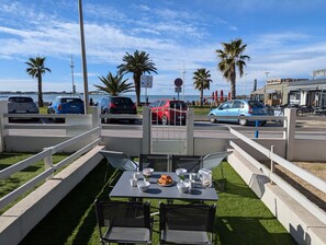 An outdoor seating area with tables and chairs, a view of the sea, and parked cars. - Sol-y-Days le Goéland, Seafront apartment with parking (Le Grau-du-Roi)