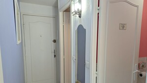 Um corredor estreito com uma porta branca à esquerda, uma porta com um espelho à direita, e uma porta com o símbolo de um sanitário no canto direito.