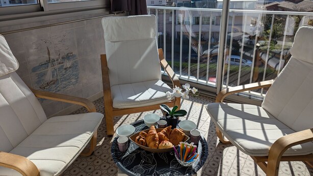 Uma varanda com duas cadeiras brancas acolchoadas, uma mesa com croissants, xícaras e uma planta.