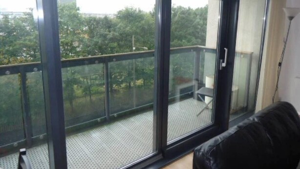 Une pièce dotée d’une grande porte-fenêtre coulissante donnant sur un balcon avec une rambarde en métal, un canapé en cuir noir et une petite table.