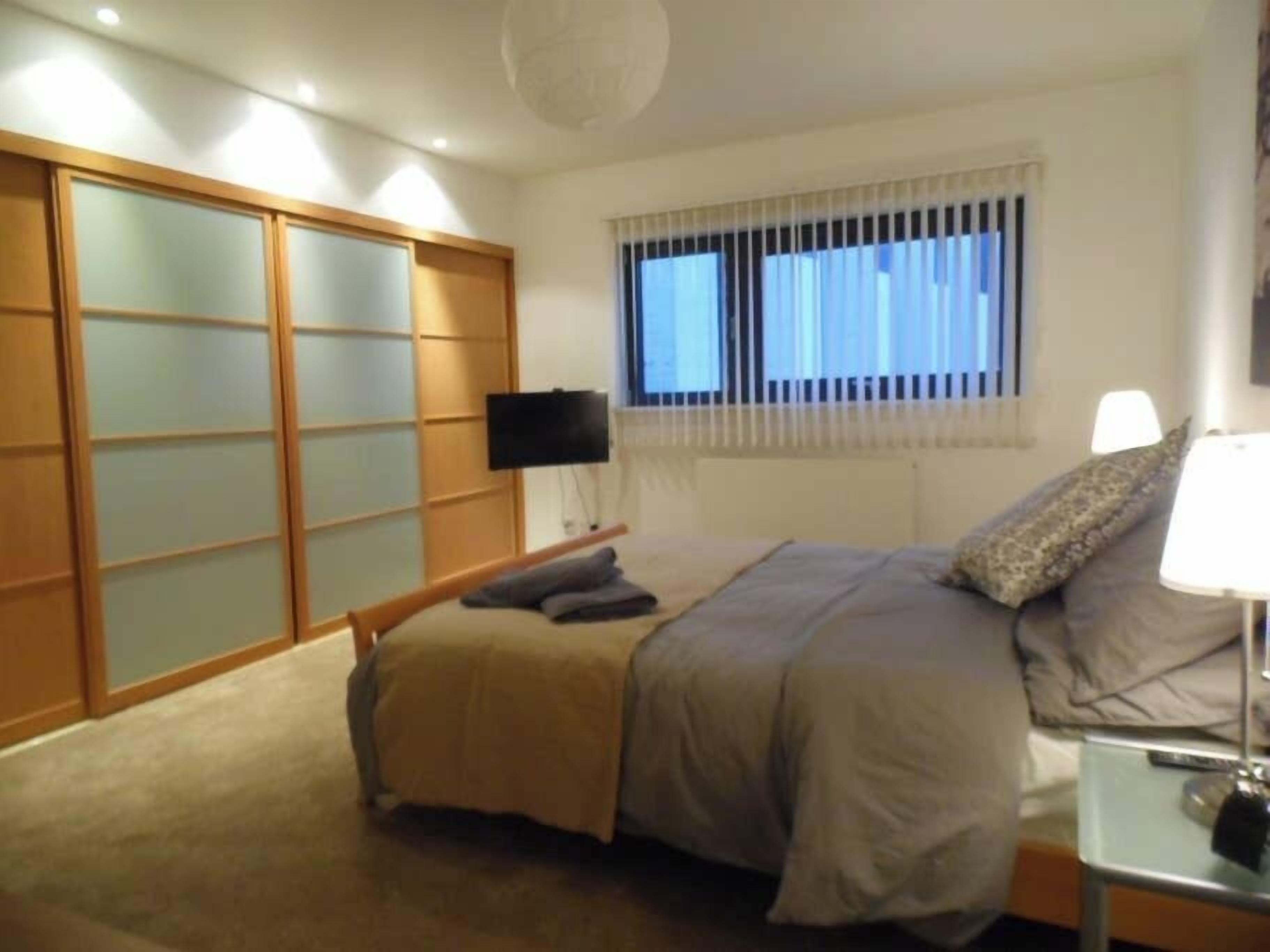 A bedroom with a bed, a bedside table with a lamp, a TV on the wall, and a window with blinds.