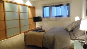 Une chambre à coucher avec un lit, une table de chevet avec une lampe, un téléviseur fixé au mur et une fenêtre avec des stores.