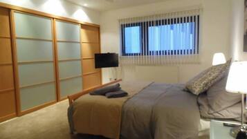 A bedroom with a bed, a bedside table with a lamp, a TV on the wall, and a window with blinds.