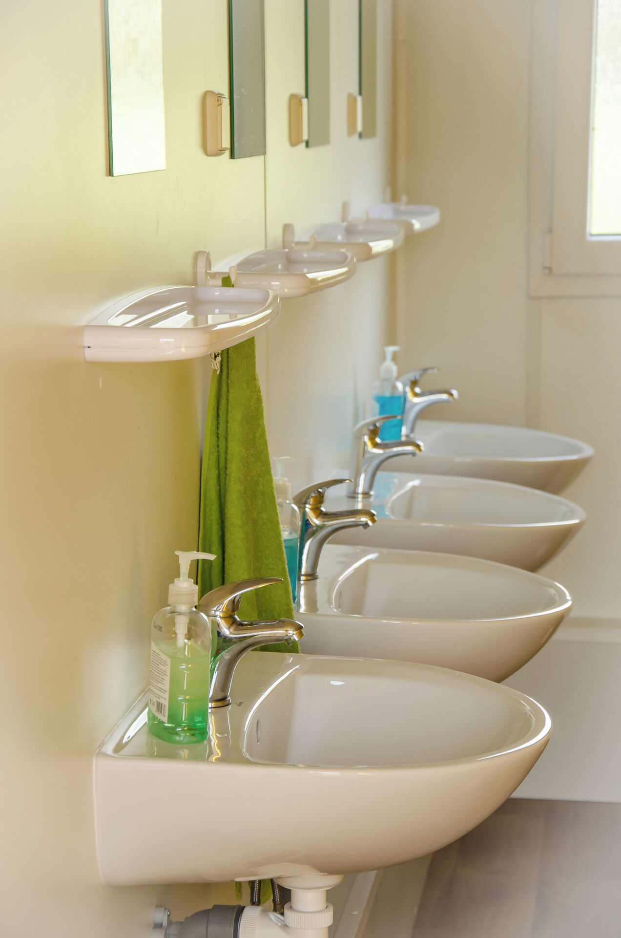 A row of white sinks with individual faucets and soap dispensers, mounted on a wall with mirrors above.