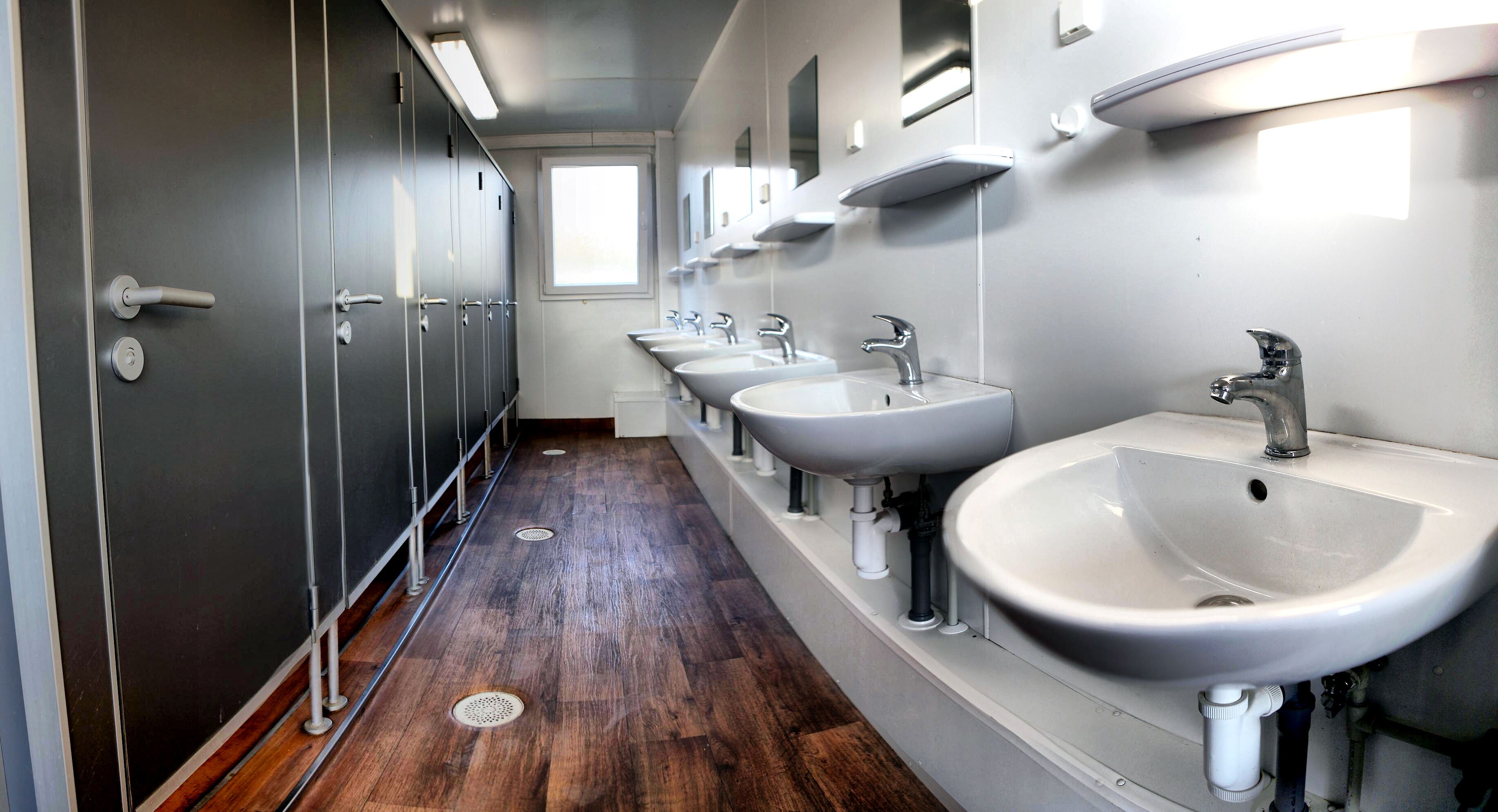 A row of sinks with mirrors above them, wooden flooring, and a window at the end of the hallway.