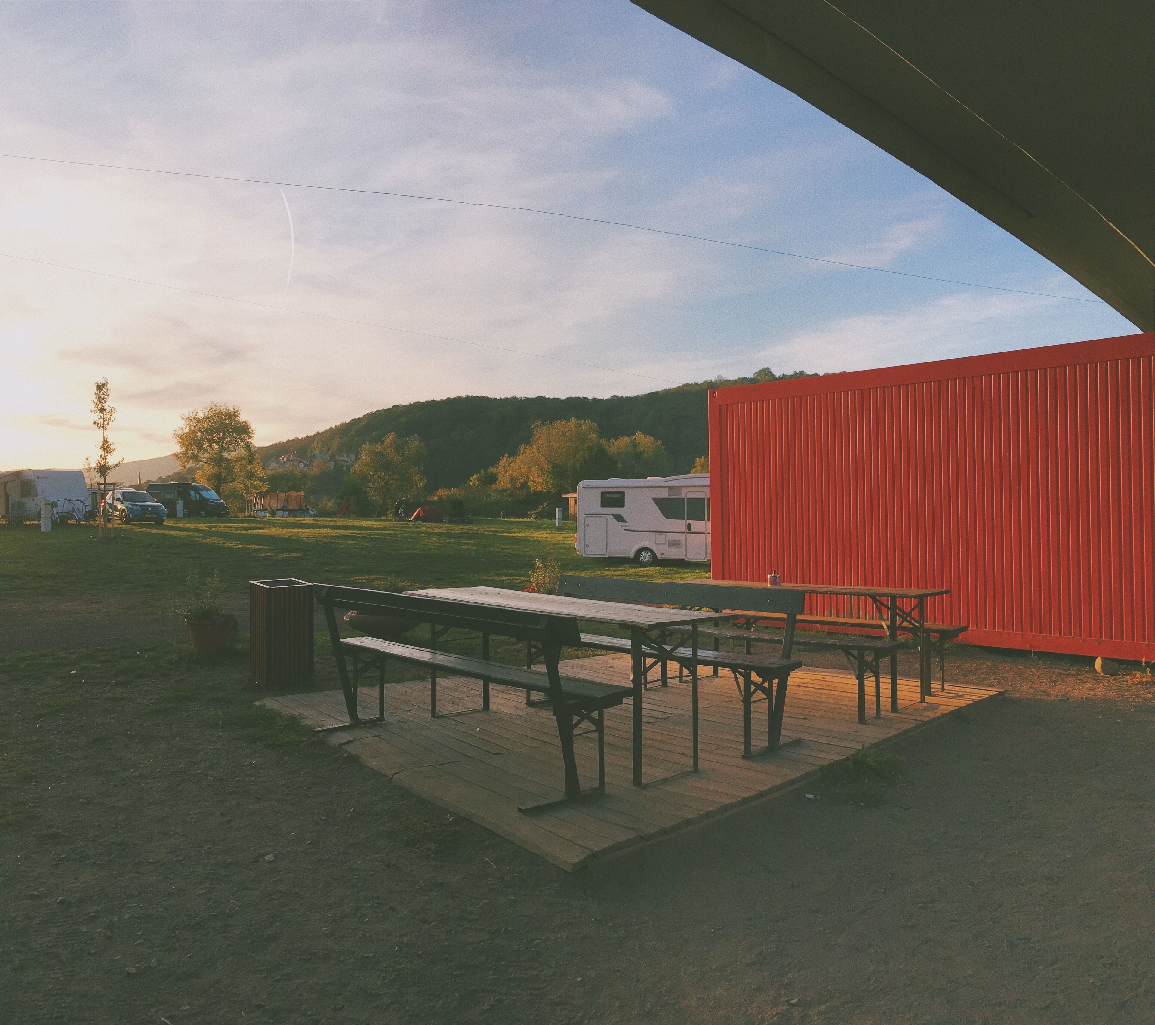 Ein Picknickbereich mit Picknicktischen und Bänken, ein rotes Gebäude und ein weißer Wohnmobil im Hintergrund.