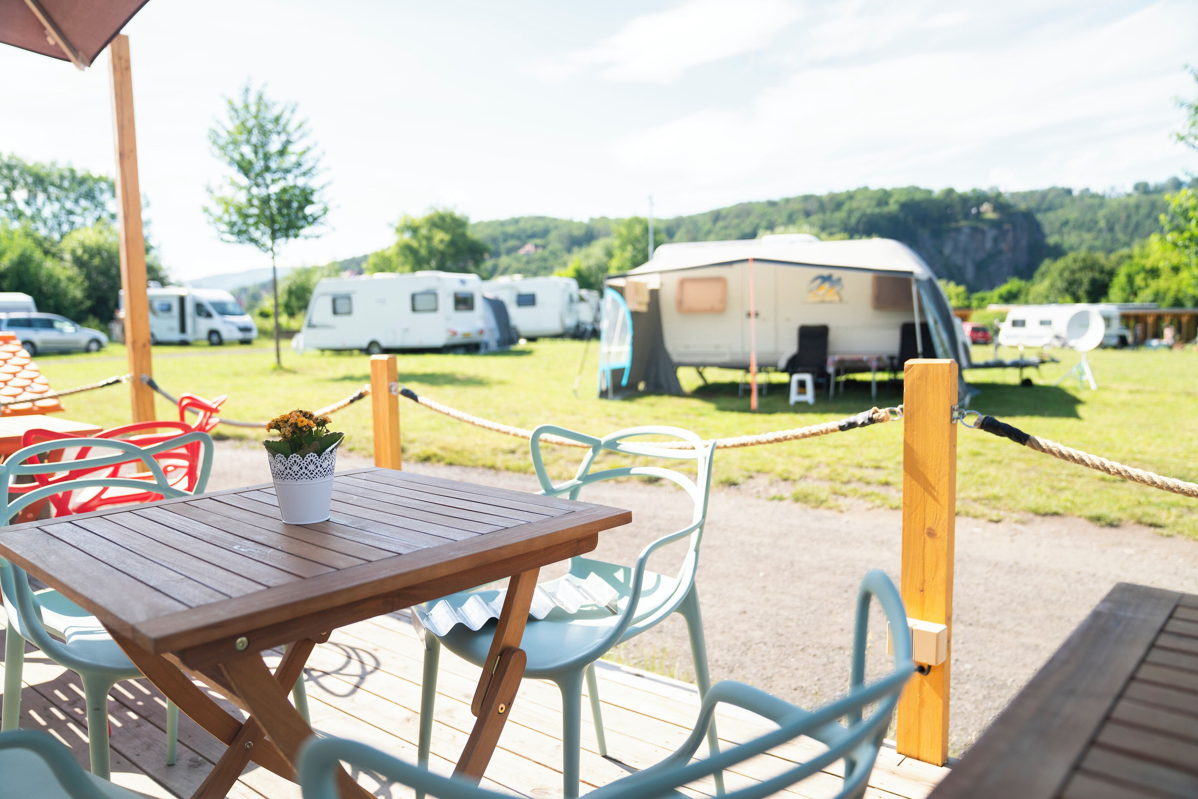Eine Holzterrasse mit Tisch und Stühlen, einer Seilsicherung und Wohnmobilen im Hintergrund.