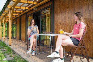 Dos personas sentadas en una mesa en una terraza al aire libre con barandas de madera y un ventanal de vidrio que da a un jardín.