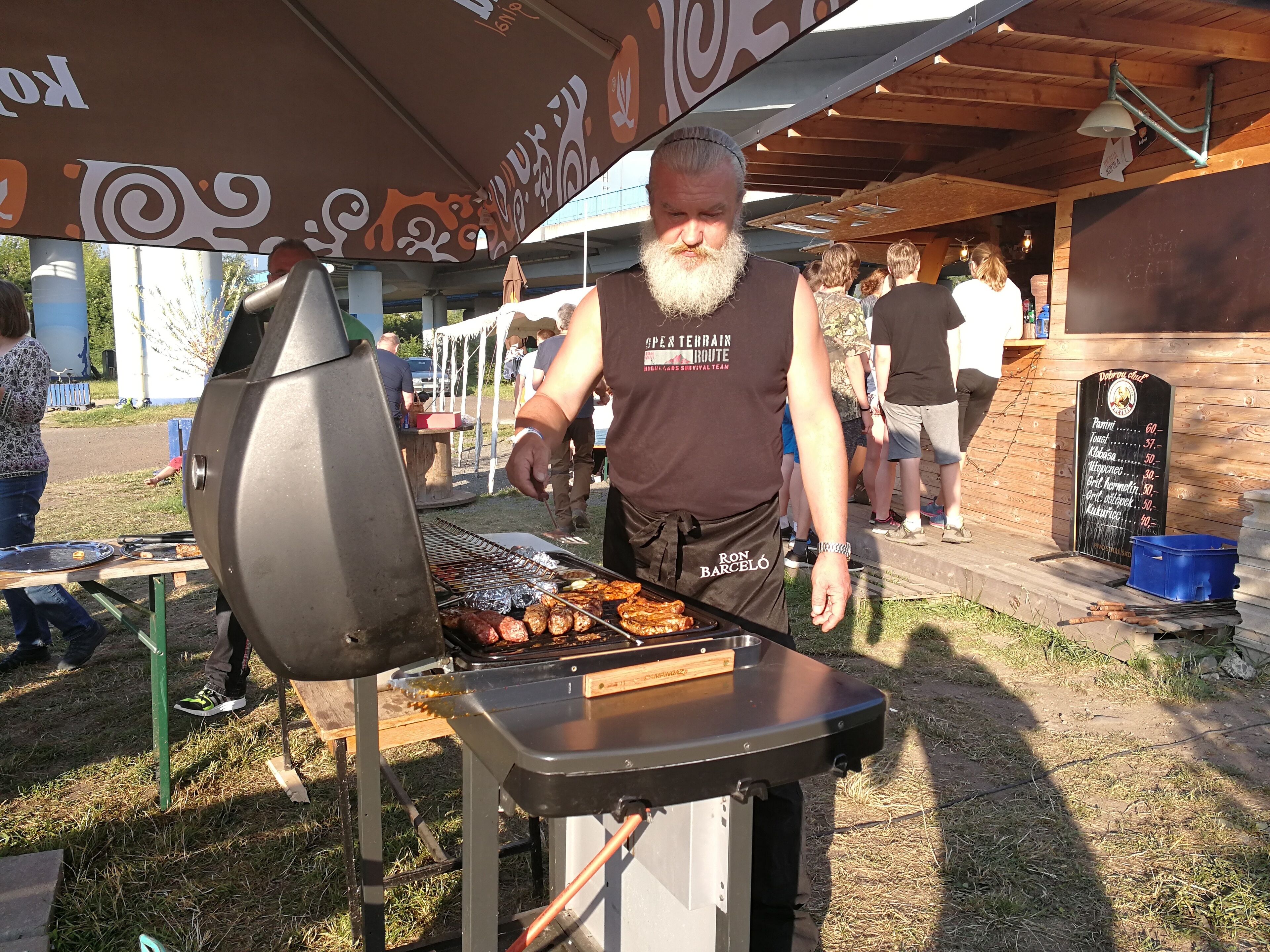 Un homme prépare des grillades lors d’un événement en plein air, et on peut voir un panneau affichant le menu en arrière-plan.