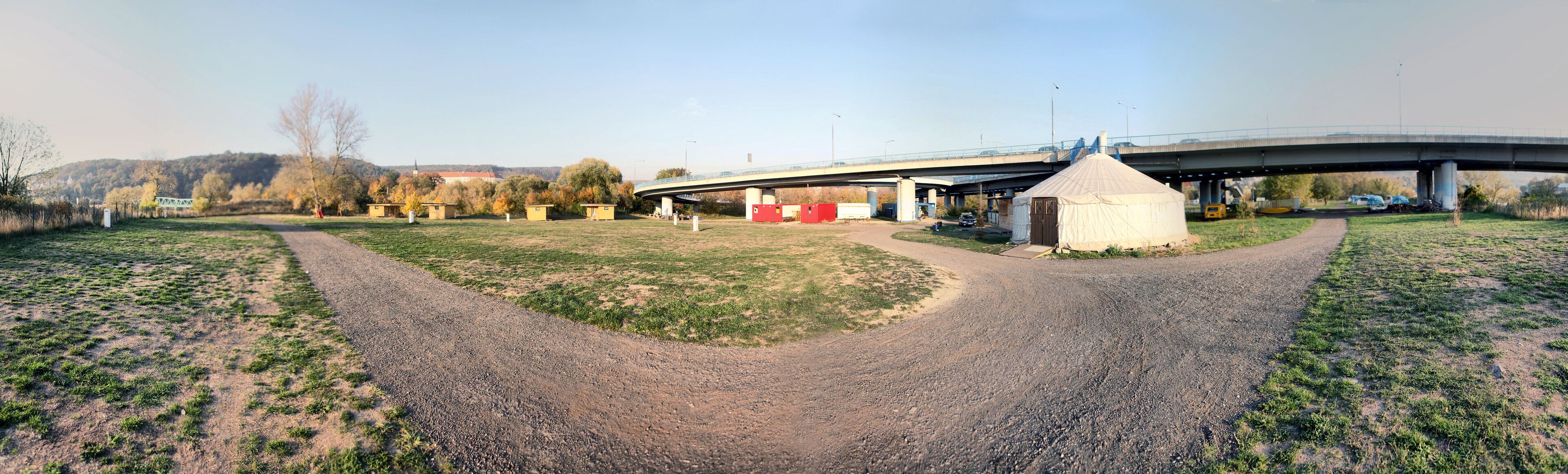 A paved path leading to a small building with a conical structure nearby, under a bridge with vehicles passing.